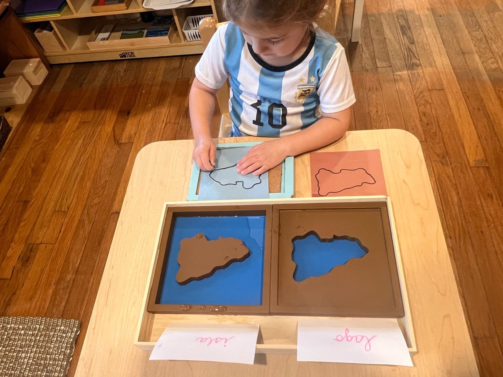 A child tracing landforms beside Montessori island and lake materials.